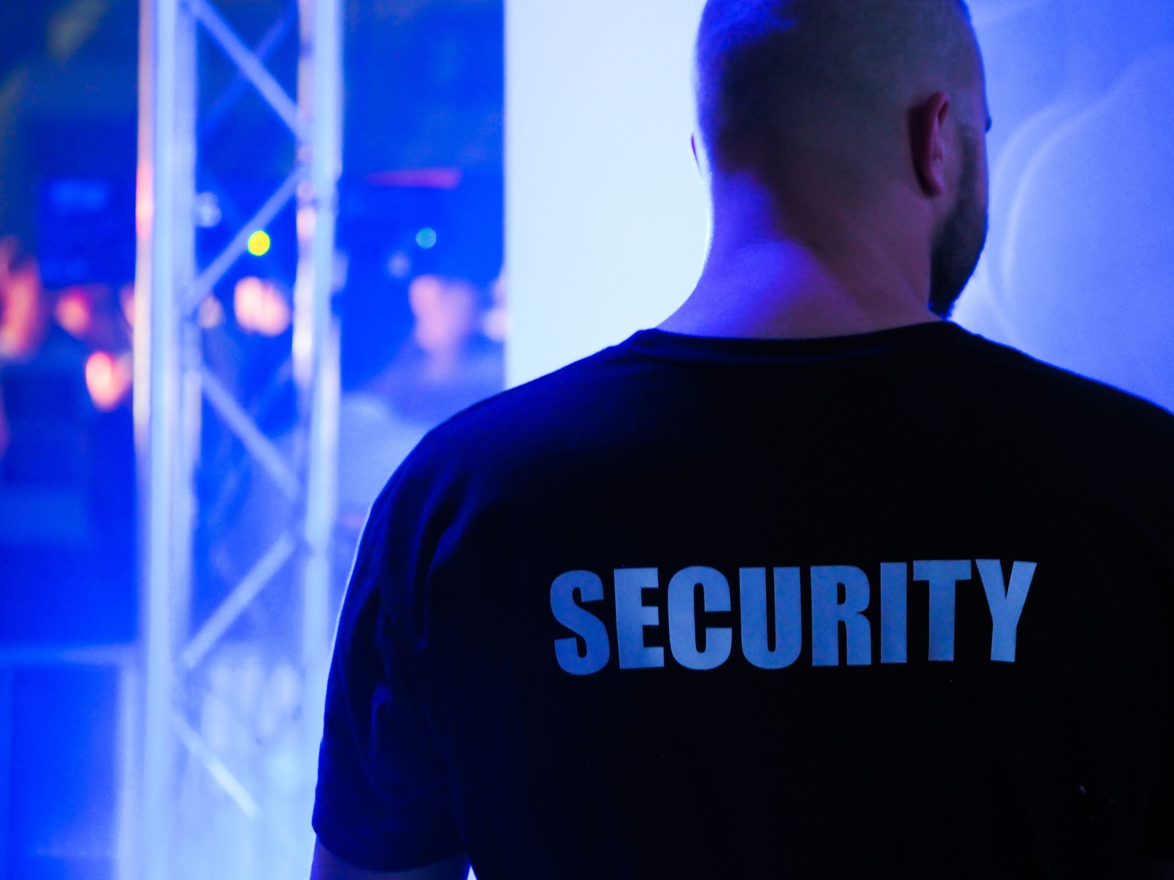 Profissional de segurança de costas, com a palavra SECURITY estampada na camisa, posicionado na entrada de um evento.