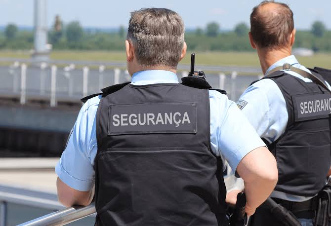 Dois profissionais de costas, com coletes escritos SEGURANÇA, observando uma área externa.